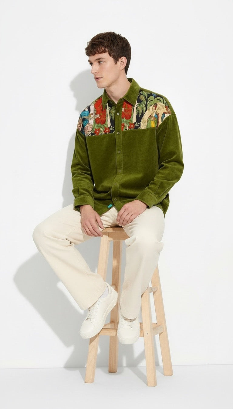 A man sits on a wooden stool wearing an olive green corduroy button-up shirt with a Frida Kahlo chest panel, cream trousers, and white sneakers against a plain white studio background.