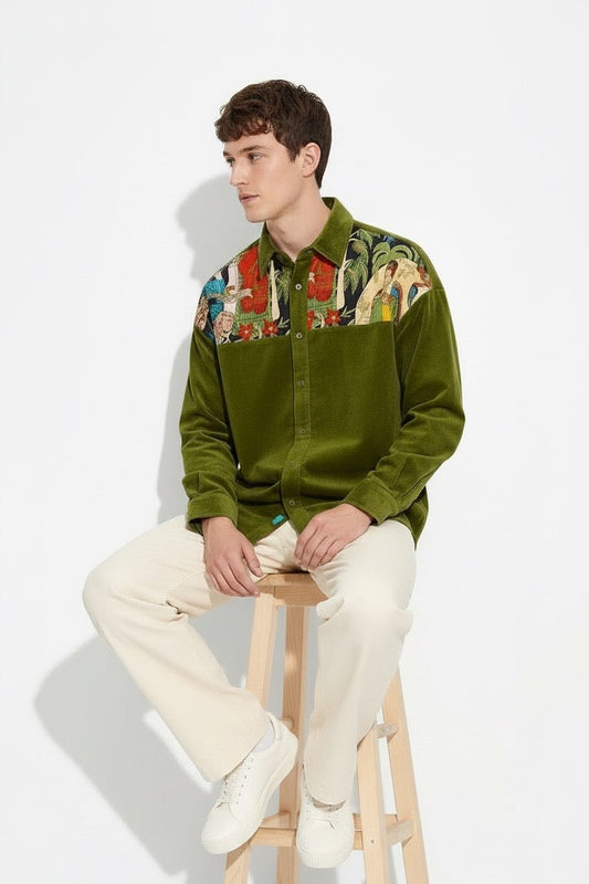 A man sits on a wooden stool wearing an olive green corduroy button-up shirt with a Frida Kahlo chest panel, cream trousers, and white sneakers against a plain white studio background.